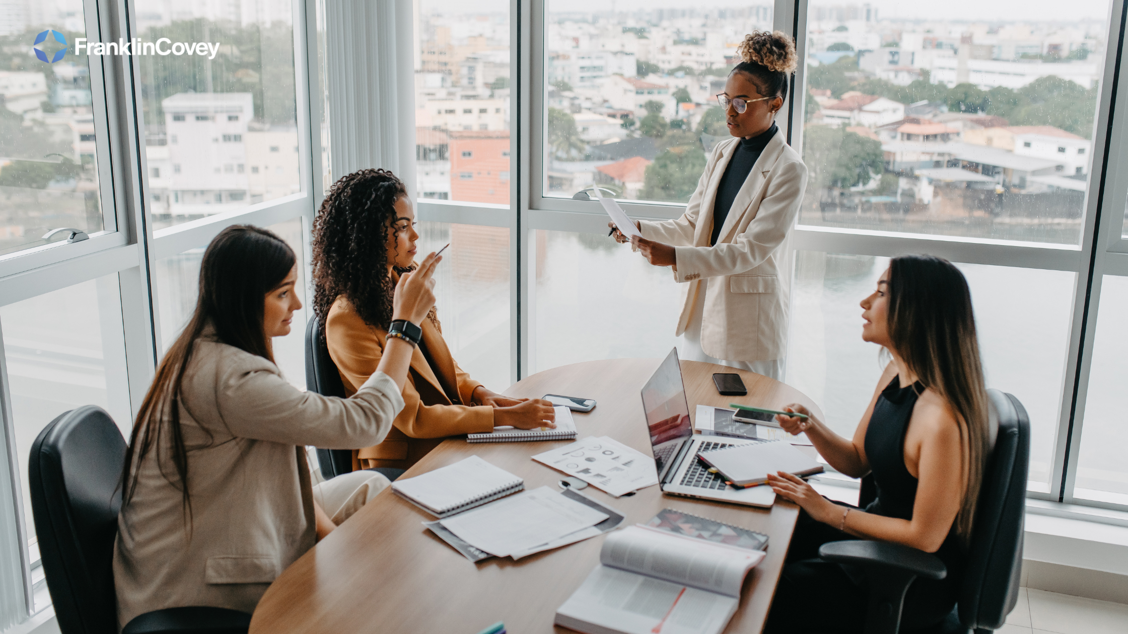 Three women who are business professionals sit round a table discussing an issue that is related to the business matter at hand, potentially discussing leadership development strategies.